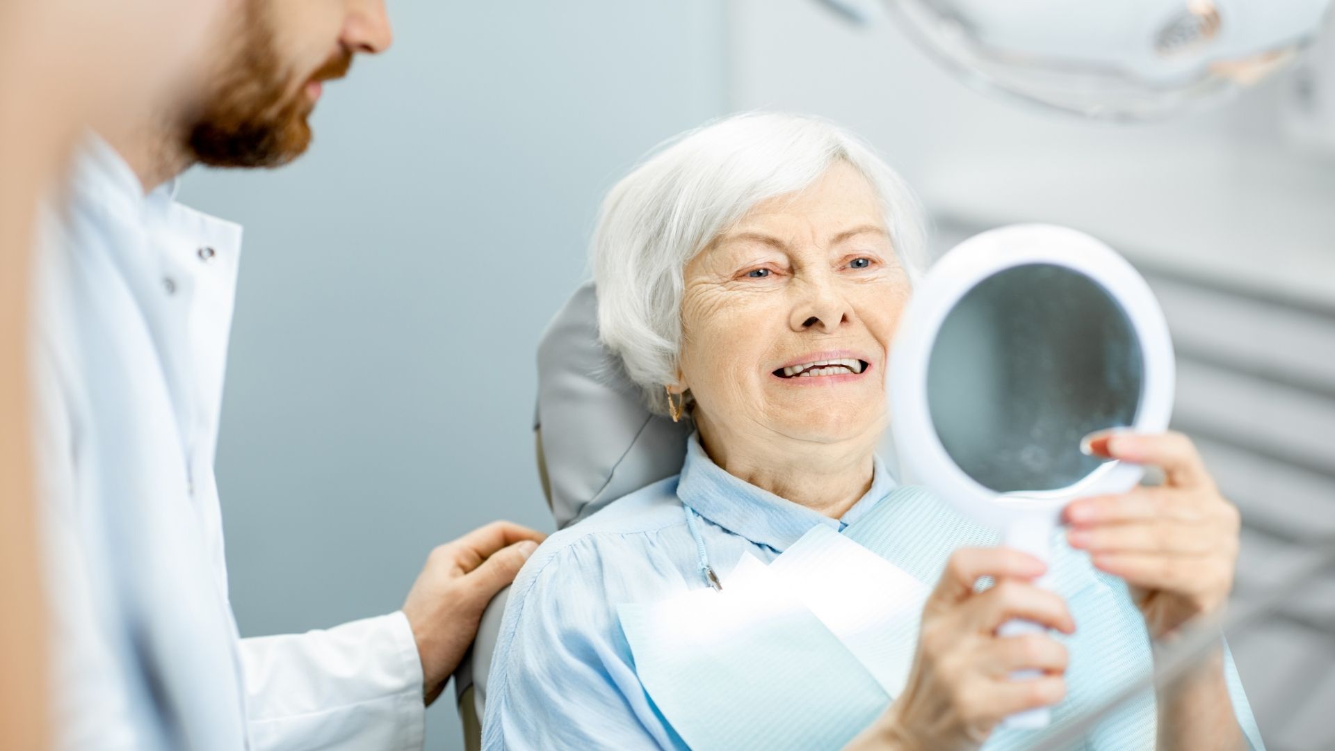 Elderly woman smiling with dentist and mirror.